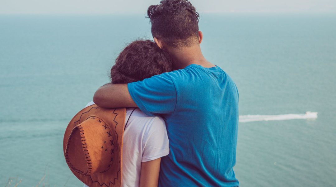 A man and a woman hugging while watching a boat sail by them