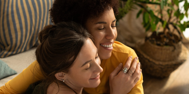 Two women hugging by a sofa and a big plant.